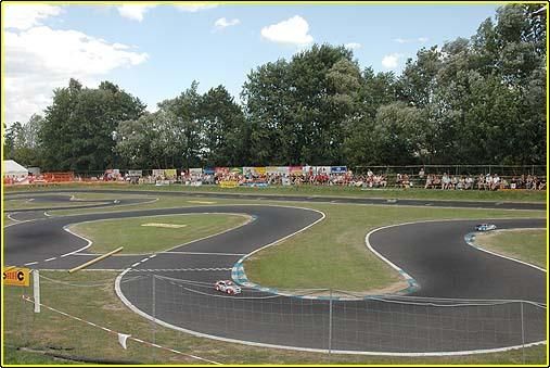 Eine Rennstrecke mit zwei Autos darauf. Ein Publikum beobachtet von den Seitenlinien. Bäume sind im Hintergrund.