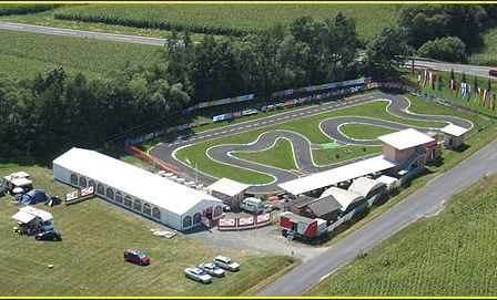 Eine Rennstrecke mit Flaggen und einer Straße mit Autos. Mehrere Zelte, Autos und Lastwagen sind im Bereich geparkt. Ein großes weißes Zelt ist links und ein Gebäude ist rechts.