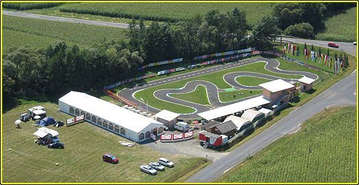 Eine Rennstrecke mit Flaggen und einer Straße mit Autos. Mehrere Zelte, Autos und Lastwagen sind im Bereich geparkt. Ein großes weißes Zelt ist links und ein Gebäude ist rechts.