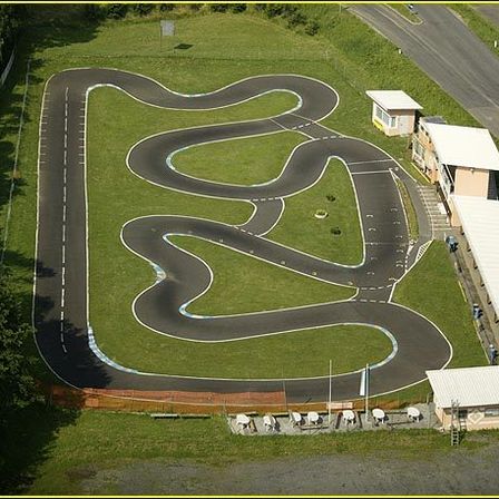 Luftaufnahme einer kurvenreichen Rennstrecke mit mehreren Gebäuden und Bäumen. Die Strecke hat weiße Markierungen und ist von einem Rasenfeld umgeben. Es gibt eine Straße, die zur Strecke führt.