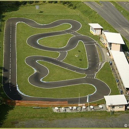 Luftaufnahme einer kurvenreichen Rennstrecke, umgeben von Rasen und Bäumen. Gebäude, eine Bushaltestelle und Fahrzeuge sind sichtbar.