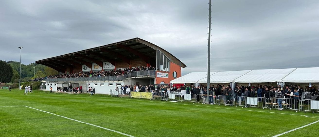 Eine große Menschenmenge versammelt sich in einem Sportstadion. Der Platz ist gut gepflegt mit weißen Markierungen. Es gibt Menschen, die hinter den Absperrungen stehen, und einige sitzen in den Rängen.