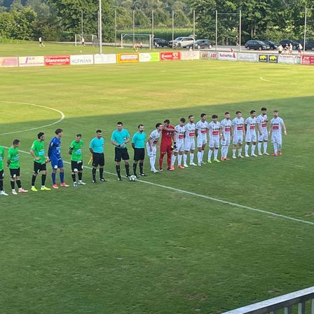 Ein Fußballspiel ist im Gange mit Spielern in grünen und weißen Trikots. Die Spieler stehen in einer Reihe und der Schiedsrichter ist in der Nähe. Das Feld hat weiße Linien und ist von einem Zaun umgeben.