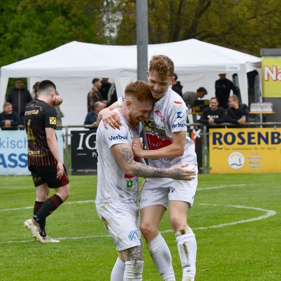 Zwei Fußballspieler umarmen sich zur Feier auf dem Spielfeld, mit weißen Zelten und einer Menge im Hintergrund.