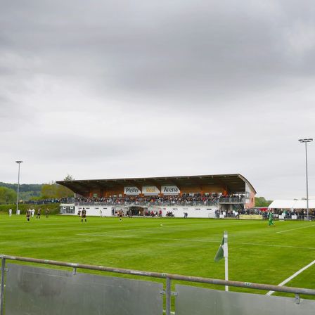 Ein Fußballfeld mit Spielern auf dem Spielfeld. Zuschauer sind in den Tribünen. Der Himmel ist bewölkt.