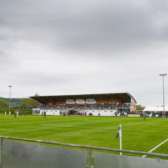 Ein Fußballfeld mit Spielern auf dem Spielfeld. Zuschauer sind in den Tribünen. Der Himmel ist bewölkt.