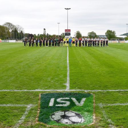 Fußballspieler stehen in zwei Reihen auf einem grünen Feld, ein Fußball-Logo ist auf dem Boden.