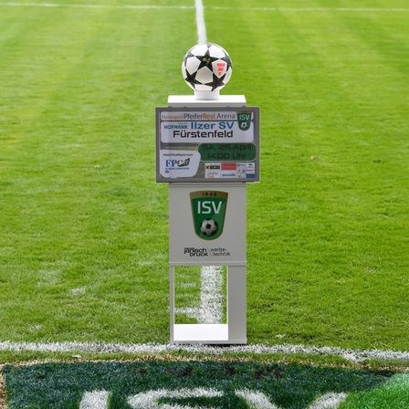 Ein Fußball steht auf einem Schild auf einem Fußballfeld. Das Schild enthält Logos, Text und Zahlen zu einem bevorstehenden Spiel.