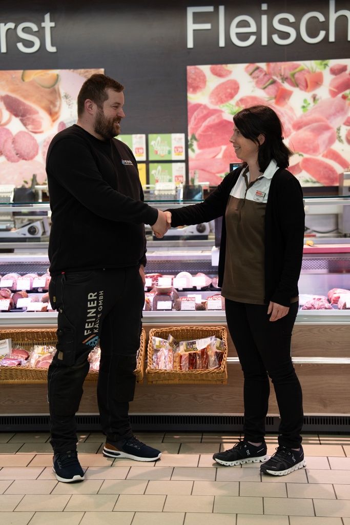 Ein Mann und eine Frau schütteln sich die Hände vor einer Fleischtheke und lächeln. Der Mann trägt ein schwarzes Shirt mit 'Feliner' und 'Kalf GmbH', während die Frau ein schwarzes und braunes Shirt trägt. Körbe mit Fleisch stehen hinter ihnen.