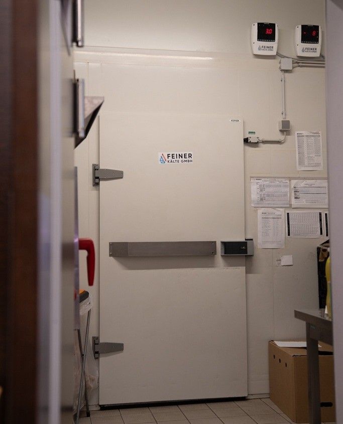 Ein Raum mit einer weißen Tür und rotem Griff, beschriftet mit Feiner Kälte GmbH. Temperaturanzeigen sind oben angebracht. Papiere sind an der Wand angeheftet.
