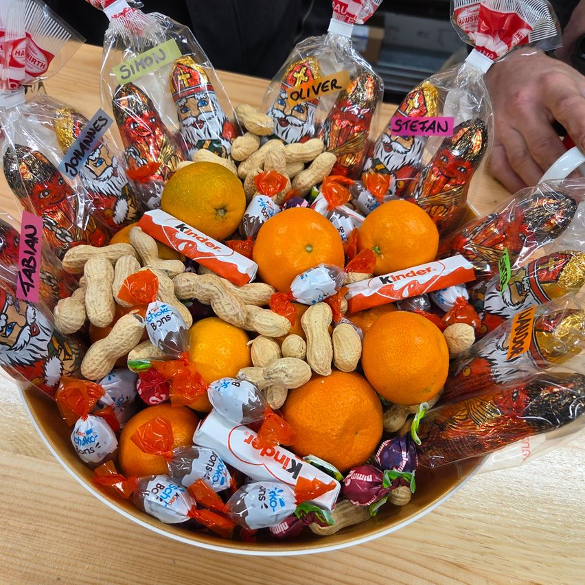 Eine Schüssel voller verschiedener Süßigkeiten, Orangen und Erdnüssen. Die Schüssel enthält Kinderschokoladen, Orangen und Erdnüsse. Einzelne Süßigkeiten sind in beschrifteten Tüten mit Namen wie Simon, Oliver und Stefan.
