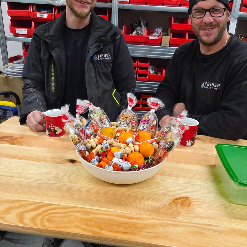 Zwei Männer in Arbeitskleidung lächeln, sitzen an einem Tisch mit einer Schüssel voller Süßigkeiten und Obst. Der Tisch hat Mickey Mouse Tassen, und sie befinden sich in einem Raum mit Regalen voller roter Aufbewahrungsboxen.