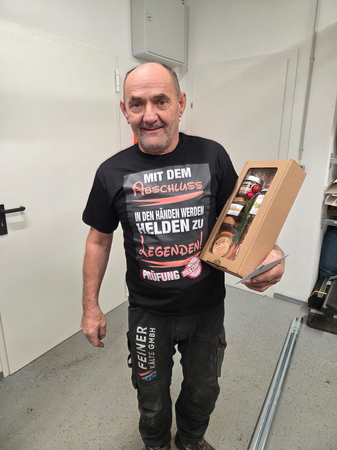 Ein Mann in einem Raum trägt ein schwarzes T-Shirt mit deutschem Text und hält eine braune Box mit Essen. Er lächelt und steht in der Nähe einer weißen Tür.