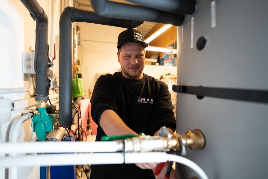 Ein Mann in einem schwarzen Shirt und einer Kappe arbeitet an einer Maschine in einem Raum mit Rohren und Werkzeugen. Er hält eine Zange und lächelt.