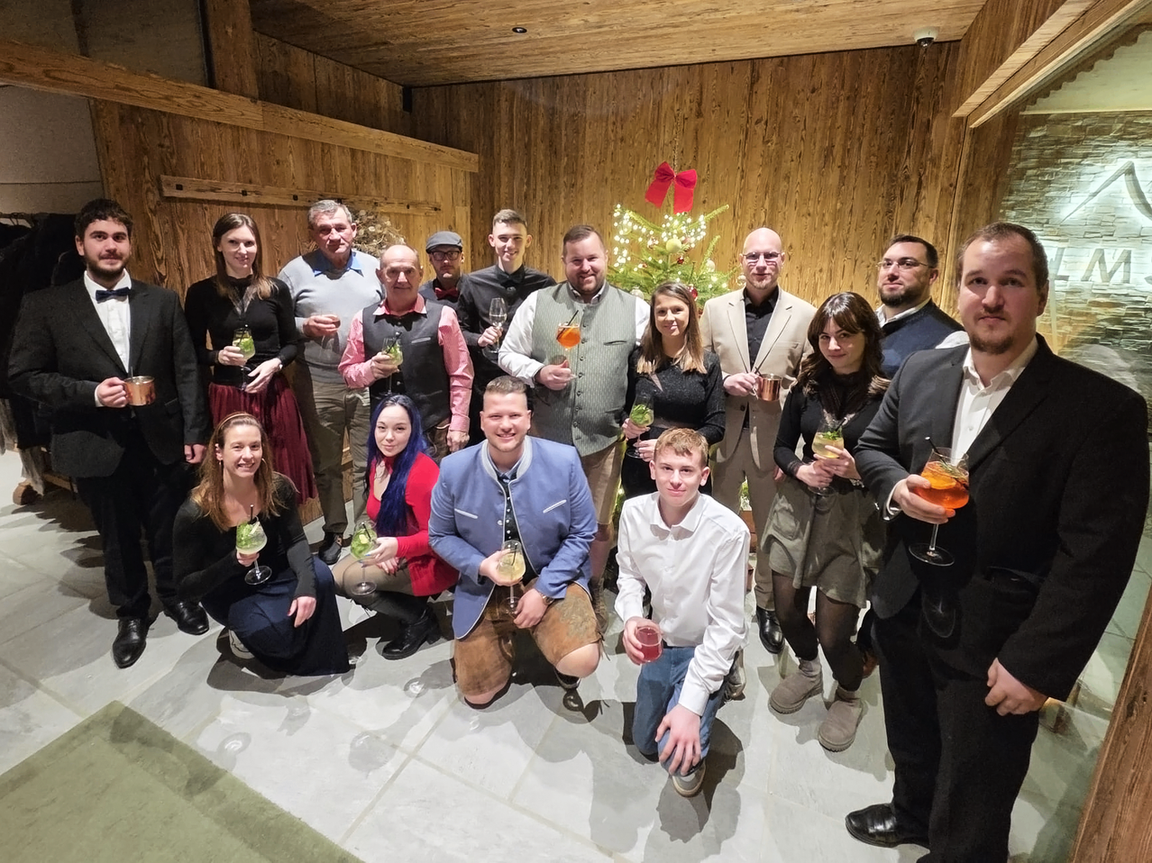 Eine Gruppe von Menschen in formeller Kleidung, einige in traditionellen Kostümen, posiert für ein Foto in einem Raum mit Holzwänden. Sie halten Getränke und lächeln. Ein Weihnachtsbaum mit rotem Band steht hinter ihnen.