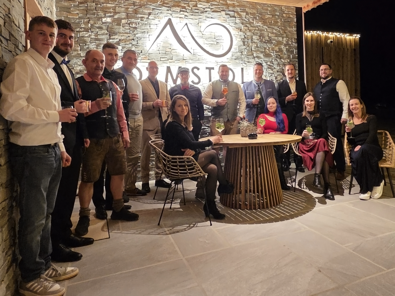 Eine Gruppe von Erwachsenen, sowohl Männer als auch Frauen, posieren für ein Foto bei einer Outdoor-Veranstaltung, halten Getränke in den Händen, mit einem Tisch in der Mitte und einer Steinmauer im Hintergrund.