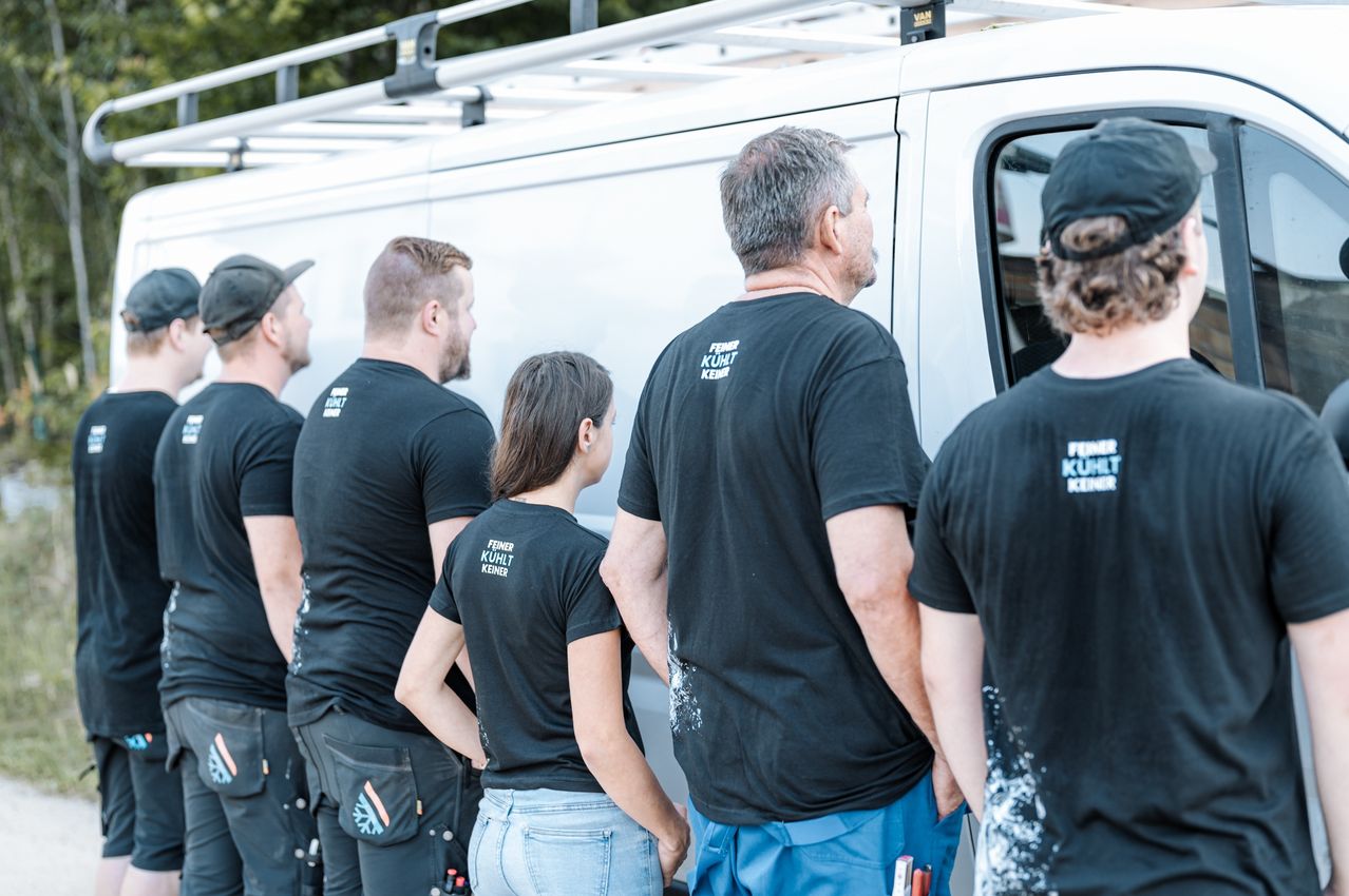 A group of six people wearing black t-shirts with white text stand in front of a white van. They are likely discussing work.