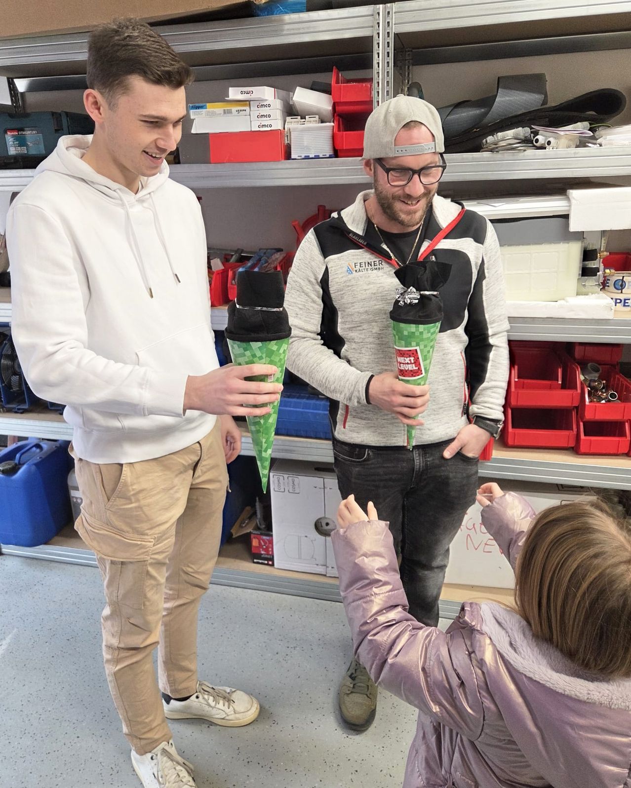 Two men in a workshop, one in a white hoodie and the other in a grey jacket, holding green cups, with a girl reaching up to them. Behind them, shelves are filled with various items.