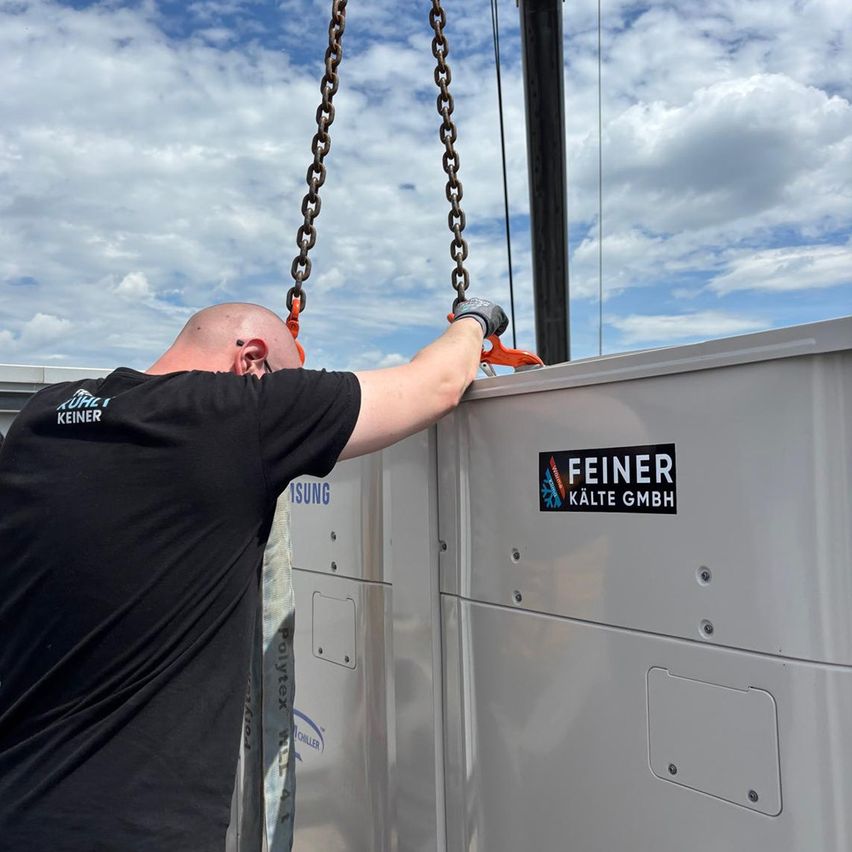 Ein Mann in einem schwarzen T-Shirt benutzt einen Kran, um eine Metallbox mit dem Logo 'Feinér Kälte GmbH' anzuheben. Der Himmel ist blau mit Wolken.