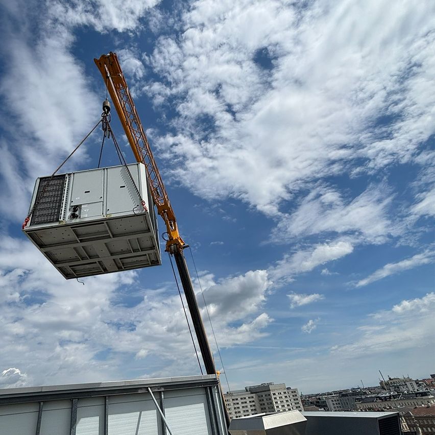 Ein Kran hebt eine große, graue Metallbox gegen einen blauen Himmel mit verstreuten Wolken, über ein Gebäude.