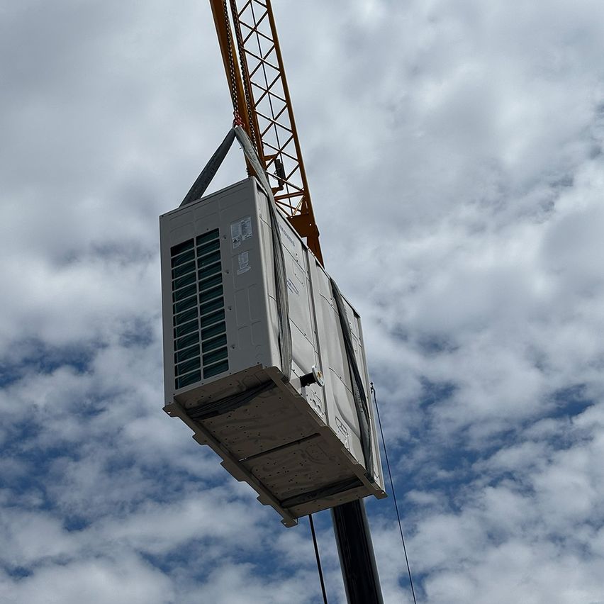 Ein Kran hebt eine große graue HVAC-Einheit mit mehreren Lufteinlässen und einem Metallrahmen gegen einen blauen Himmel mit Wolken an.
