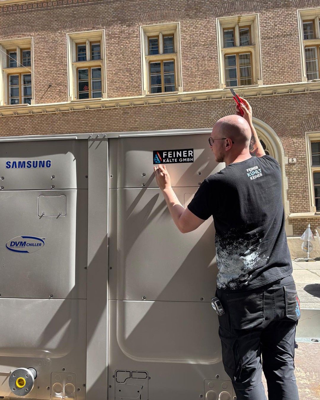Ein Mann steht vor einer Metallstruktur und hält Scheren. Die Struktur hat Samsung- und DVM-Kühler-Logos. Er trägt ein schwarzes T-Shirt mit einem weißen Logo. Dahinter steht ein Ziegelgebäude.