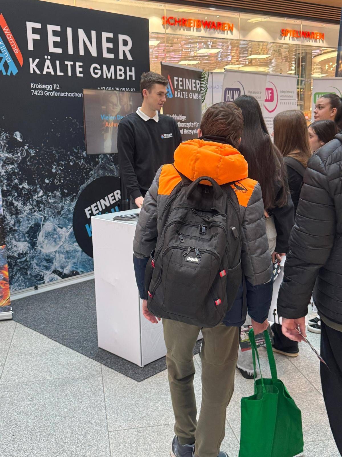 A group of people stand in a line at a booth with a large banner behind it. A man speaks to a young person with a backpack.
