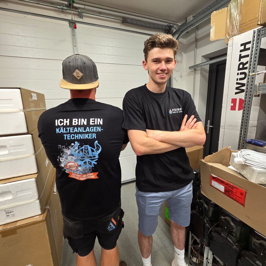 Zwei Männer in einem Lager mit Lagerboxen. Einer trägt ein schwarzes T-Shirt mit deutschem Text und der andere lächelt mit verschränkten Armen.
