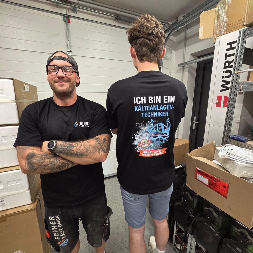Zwei Männer in einem Lagerraum, einer mit Tattoos und einer Kappe, der andere mit einem Shirt, auf dem steht 'Ich bin ein Kälteanlagen-Techniker.' Hinter ihnen stehen Kisten und ein Wurth-Schild.