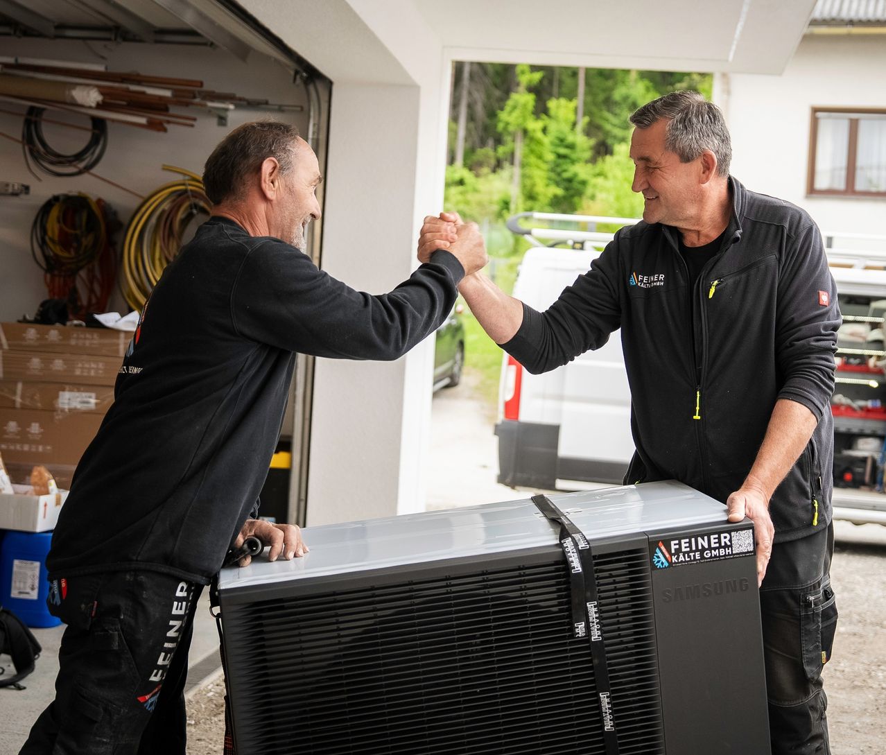 Two men are shaking hands near a garage door, one holding a Samsung appliance, with a vehicle and trees in the background.