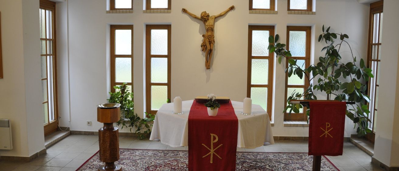 Ein Altar in einer Kirche mit einem Kreuz an der Wand, geschmückt mit einem roten Tuch, das ein goldenes 'P' zeigt. Zwei Kerzen und eine Blume in einem Topf sind auf dem Altar platziert. Es gibt sechs Fenster an der Wand und zwei Topfpflanzen auf beiden Seiten des Altars.