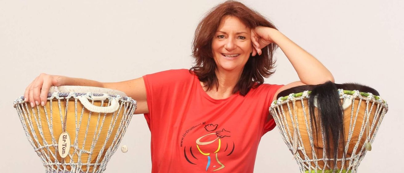 Eine Frau in einem roten T-Shirt mit einem grafischen Design sitzt zwischen zwei Djembe-Trommeln auf dem Boden. Sie hat die Hand auf dem Kopf und lächelt.