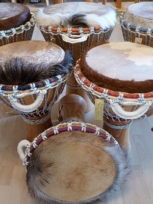 Mehrere hölzerne Trommeln mit Fell- und Seilverzierungen sind auf einem Holzboden angeordnet. Die Trommeln variieren in Größe und Form, und einige haben angebrachte Etiketten.