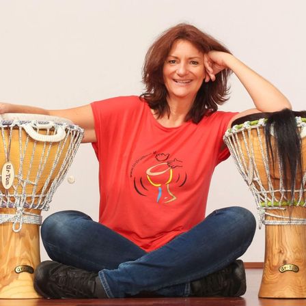 Eine Frau sitzt mit verschränkten Beinen mit zwei Trommeln, eine mit der Aufschrift 'Djembe' und die andere 'Djembe'. Sie trägt ein rotes T-Shirt mit einem Trommel-Grafik und blaue Jeans. Ihr Haar ruht auf der Trommel mit der Aufschrift 'Djembe'.