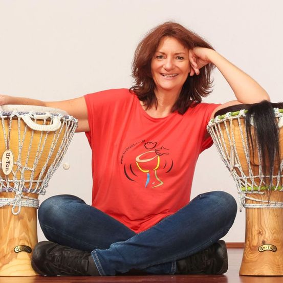 Eine Frau sitzt mit verschränkten Beinen mit zwei Trommeln, eine mit der Aufschrift 'Djembe' und die andere 'Djembe'. Sie trägt ein rotes T-Shirt mit einem Trommel-Grafik und blaue Jeans. Ihr Haar ruht auf der Trommel mit der Aufschrift 'Djembe'.