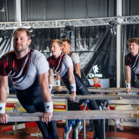 Vier Männer in passenden Uniformen führen Gymnastikübungen an Parallelbarren in einer Turnhalle durch. Sie lächeln und scheinen mitten in einer Routine zu sein.