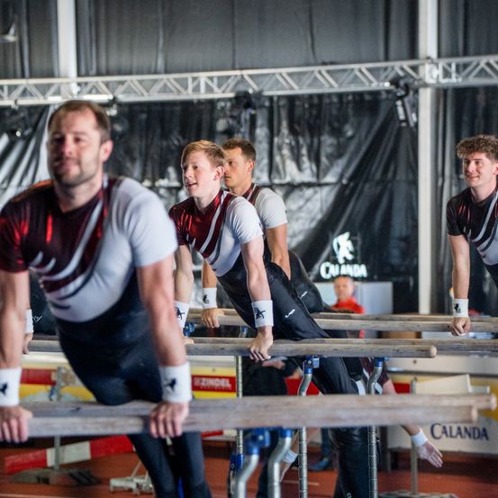 Vier Männer in passenden Uniformen führen Gymnastikübungen an Parallelbarren in einer Turnhalle durch. Sie lächeln und scheinen mitten in einer Routine zu sein.