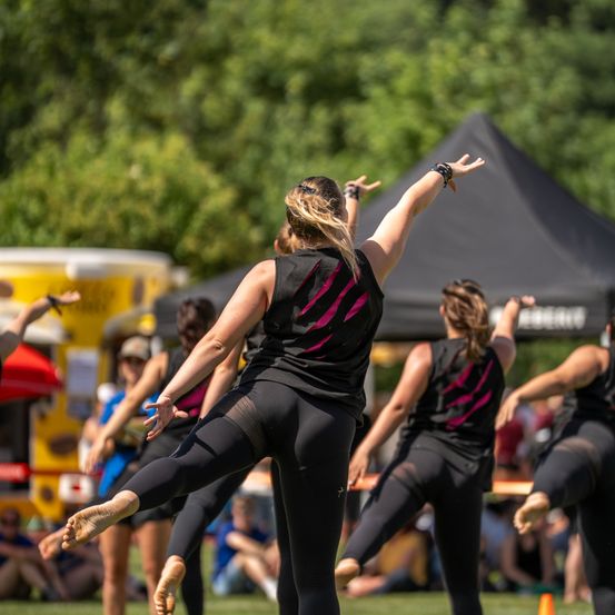 Eine Gruppe von Frauen in passenden schwarzen und pinkfarbenen Uniformen führt einen synchronisierten Tanz auf einem Rasenfeld mit einem schwarzen Zelt im Hintergrund auf.