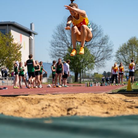 Eine Athletin in einem gelben und grünen Trikot springt über eine Sandgrube bei einem Leichtathletik-Event. Zuschauer und andere Athleten sind im Hintergrund zu sehen.