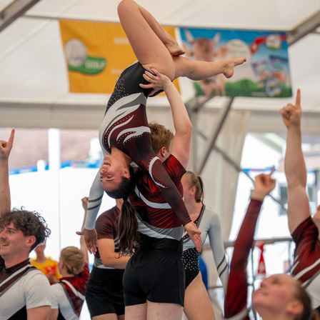 Eine Turnerin führt einen Handstand aus, während sie von einer anderen Turnerin gestützt wird, in einer Turnhalle, mit jubelnden Zuschauern im Hintergrund.