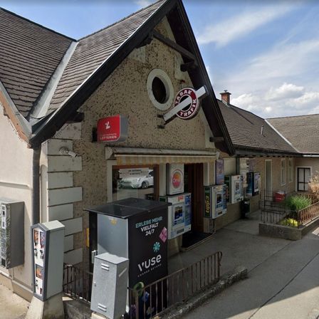 Ein Laden an einer Straßenecke mit einem Schild, das 'TABAK TRAIL' über dem Eingang zeigt. Es hat einen 'Vuse' Ladestationsaußen.