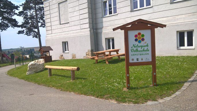Ein Holzschild steht auf einem Rasen in der Nähe eines weißen Gebäudes mit mehreren Fenstern. Ein Picknicktisch und eine Bank sind auf dem Rasen aufgestellt. Bäume und ein großer Stein befinden sich in der Nähe.
