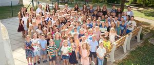 Eine große Gruppe von Kindern und Erwachsenen posiert für ein Foto im Freien, mit einer Holzbank, einem Baum und einem Spielplatz im Hintergrund.