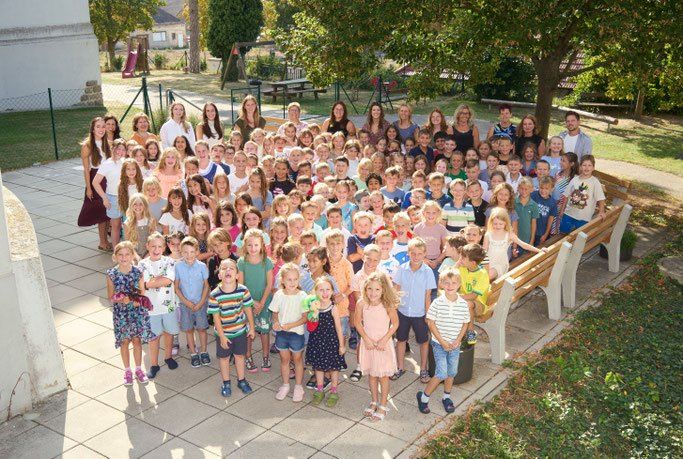 Eine große Gruppe von Kindern und Erwachsenen posiert für ein Foto im Freien, mit einer Holzbank, einem Baum und einem Spielplatz im Hintergrund.
