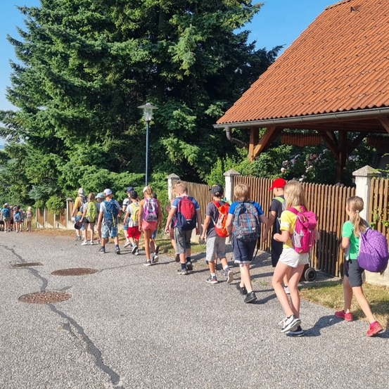 Eine Gruppe von Menschen geht mit Rucksäcken auf einem Weg, in der Nähe eines Hauses mit braunem Dach und einer Holzzaun. Bäume sind im Hintergrund.