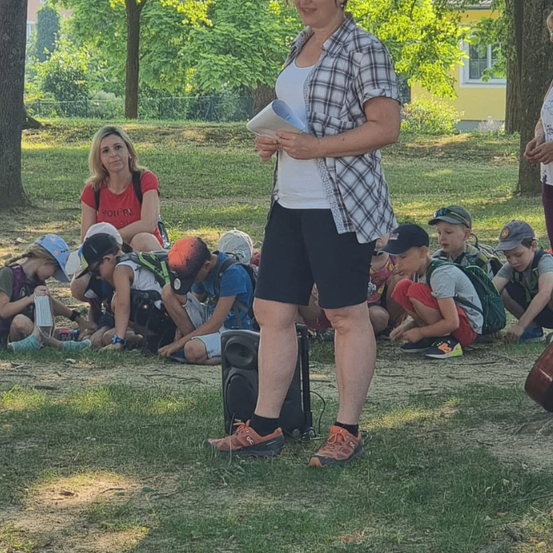 Eine Frau steht und hält ein Buch in der Hand, während eine Gruppe von Kindern vor ihr auf dem Gras sitzt. Die Kinder tragen Mützen und Turnschuhe. Bäume und ein Gebäude sind im Hintergrund.