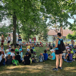 Eine Gruppe von Kindern und Erwachsenen sitzt im Kreis unter einem Baum. Eine Erwachsene steht in der Mitte. Gebäude und weitere Bäume sind im Hintergrund.