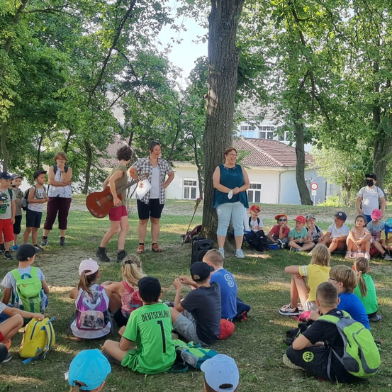 Eine Gruppe von Kindern und Erwachsenen sitzt und steht in einem Park. Einige halten Gitarren. Zwei Frauen stehen vor den Kindern. Bäume und ein Gebäude sind im Hintergrund.