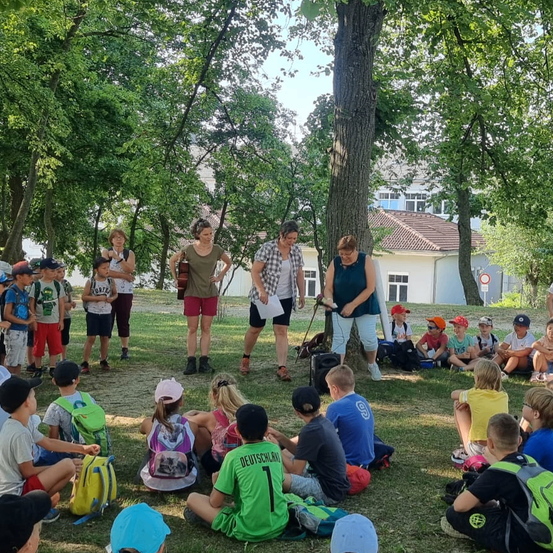 Eine Gruppe von Kindern und Erwachsenen versammelt sich in einem Park. Mehrere Kinder sitzen auf dem Gras, während andere stehen. Eine Erwachsene hält eine Gitarre. Eine Frau in einem weißen Shirt hält einen Stock. Ein Gebäude ist im Hintergrund.