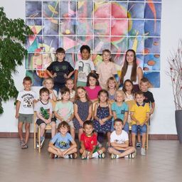 Eine Gruppe von Kindern posiert für ein Foto in einem Klassenzimmer mit einer farbenfrohen Mosaikwand, Topfpflanzen und dekorativen Zweigen auf beiden Seiten.